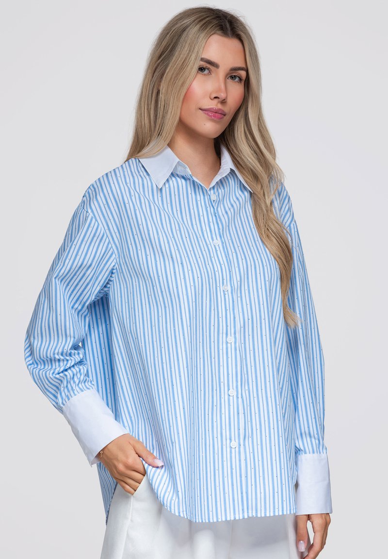 Femme aux longs cheveux blonds portant une chemise à rayures verticales bleues et blanches avec col et poignets blancs, debout avec une main dans la poche.