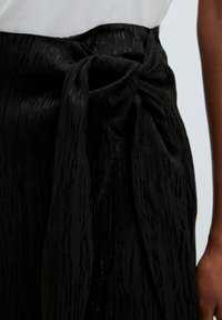 Zwarte stoffen riem met een gestructureerd patroon, voorzien van een knoopdesign en franjes. Het materiaal lijkt glad met een lichte glans.