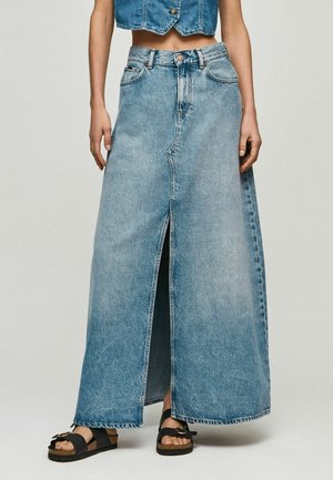 Kvinde iført lang højhalset denimnederdel med frontsplit og sorte sandaler med spænder mod en ensfarvet baggrund.