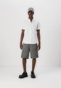 Weißes kurzärmliges Hemd, graue Cargo-Shorts und schwarze klobige Schuhe. Die Kappe ist grau mit bunten gestickten Akzenten.