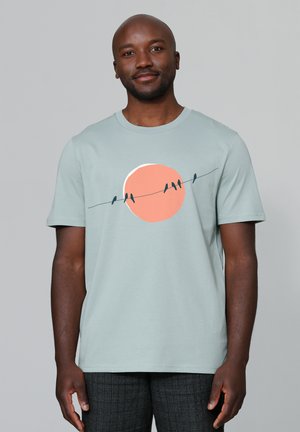 Muntgroene T-shirt met korte mouwen, voorzien van een grafisch ontwerp van vogels op een draad voor een perzikkleurige zon. Zacht katoenen materiaal, ontspannen pasvorm.