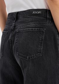 Zwarte spijkerbroek van denim met een gladde textuur, voorzien van een merkband en twee achterzakken. Gestikte accenten benadrukken de ontwerpdetails.