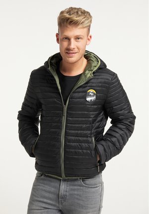 Homme portant une veste matelassée noire avec une doublure de capuche verte et un jean gris, mains dans les poches, souriant, sur un fond clair uni.