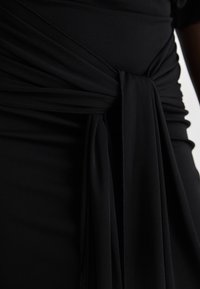 Abito nero con dettaglio annodato, texture morbida e tessuto arricciato, che crea pieghe e un movimento delicato attorno alla vita.