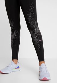 Svarta träningsleggings med ett fläckigt mönster i vitt och lila, med en Nike-logotyp på underbenet, i kombination med ljusa sneakers.