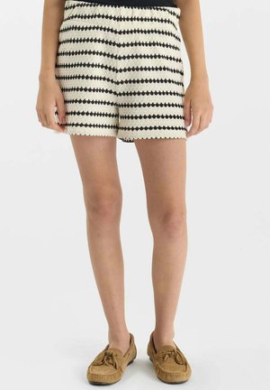 Femme portant un short en tricot rayé noir et blanc et des mocassins en daim marron, debout sur fond blanc.