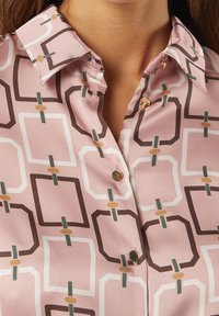 Pink bluse med et geometrisk mønster i hvid, brun og grøn. Den har en krave og knapper i guldfarve. Glat tekstur.