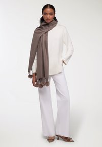 Sciarpa marrone con accenti a pom-pom drappeggiata su un maglione bianco, abbinata a pantaloni bianchi a campana e scarpe con tacco marroni. Texture lisce in tutto.