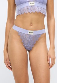 Modrček iz čipke sivke z valovitim robom, z širokim elastičnim pasom, označenim z napisom "HUGO" v kontrastni barvi. Nežni detajli cvetličnega vzorca.