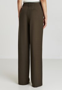 Femme portant un pantalon marron taille haute et large, se tenant avec les mains détendues sur les côtés devant un fond uni.
