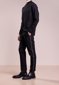 Svart långärmad skjorta med mandarin-krage, matchad med svarta byxor med vita sidostråk, och polerade svarta ankelskora.