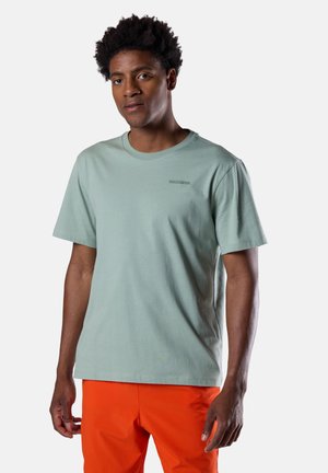 Uomo con capelli corti e ricci che indossa una maglietta a maniche corte verde chiaro e pantaloni arancioni vivaci, in piedi contro uno sfondo chiaro e uniforme.