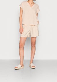 Top beige a maniche corte con scollatura a V; tessuto leggero. Abbinato a pantaloni corti beige chiaro con pieghe e tasche laterali. Sandali slip-on neutri.
