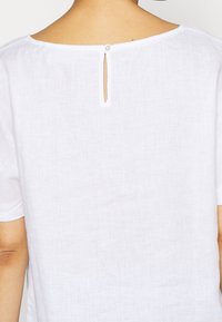 Valkoinen pellava-toppi, jossa on pyöristetty kaula-aukko ja takaosa halkio, joka on kiinnitetty pienellä nappilla. Lyhyet hihat ja pehmeä, teksturoidusta kankaasta valmistettu. 