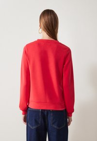 Sudadera roja de manga larga y cuello redondo, con una textura suave y un dobladillo entallado. Llevada con jeans de mezclilla azul oscuro.