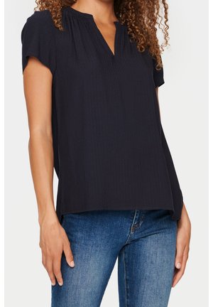 Femme portant un chemisier noir à manches courtes avec un col en V et un jean bleu en denim, montrée du cou aux hanches sur un fond uni.