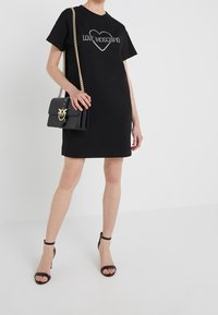 Černé šaty ve tvaru trička s designem „LOVE MOSCHINO“ z kamínků. Spárováno s černou kabelkou přes rameno se zlatým emblémem a popruhy. Černé sandály na podpatku.