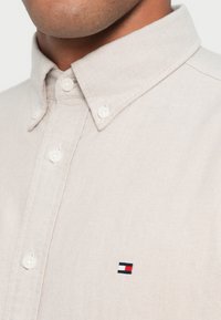 Béžová košeľa zapínaná na gombíky s klasickým golierom, s malým vyšívaným logom v námorníckej modrej, červenej a bielej na ľavej strane hrude. Hladká textúra.