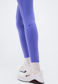 Μωβ αθλητικό κολάν με εφαρμοστή σχεδίαση, με μαύρο λογότυπο της Nike στο πλάι. Συνδυάζεται με λευκά αθλητικά παπούτσια και ανοιχτογκρίζες κάλτσες.