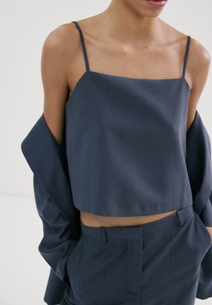 Mujer con camisola corta azul marino y pantalones de talle alto a juego, con un blazer suelto colgado en los brazos.