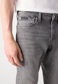 Jeans de mezclilla gris con corte ajustado, que presentan un diseño clásico de cinco bolsillos, herrajes en tono plateado y una etiqueta negra de Calvin Klein en la cintura.