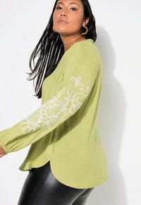 Top a maniche lunghe verde lime con ricami floreali bianchi sulla manica sinistra, caratterizzato da un orlo curvo e una vestibilità rilassata. Tessuto morbido.