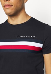 Mustat puuvillapaita, jossa on pyöreä kaulus, vaakasuorat raidat punaisena, valkoisena ja sinisenä, ja "TOMMY HILFIGER" -logo painettuna valkoisena.