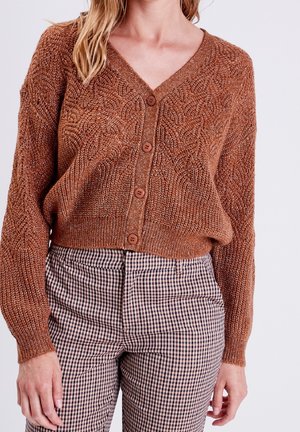 Femme portant un cardigan en maille texturée marron avec des boutons et un pantalon taille haute à carreaux dans des tons neutres.