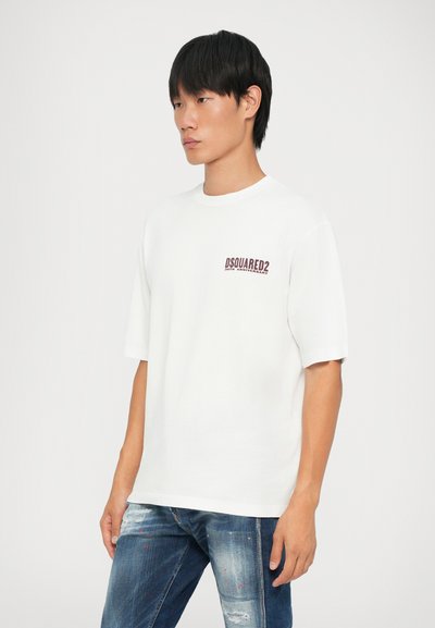 Camiseta blanca de manga corta hecha de algodón, con un estampado de logo burdeos en el lado izquierdo del pecho. Combinada con unos vaqueros azules desgastados.
