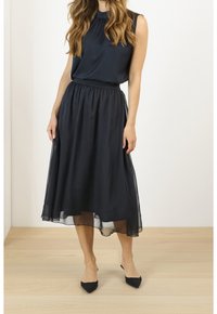 Žena ve tmavě modrém tílku a průsvitné černé midi sukni stojící na světlé dřevěné podlaze před bílým pozadím.