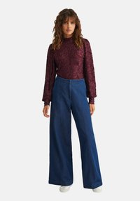 Top lung cu maneci lungi de culoare burgundy cu textura, asortat cu blugi albastri cu talie inalta si fason evazat; model purtand adidasi albi.