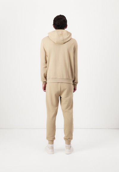 Sweat à capuche beige et pantalon de survêtement assorti en tissu doux, avec une capuche à cordon et des poignets côtelés. Porté avec des baskets claires.