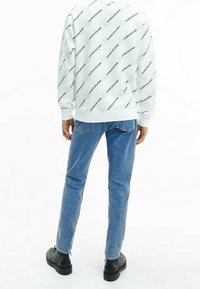 Felpa bianca con motivo ripetuto di logo nero, abbinata a jeans in denim blu e stivali neri. Forma slim-fit e polsini a costine.