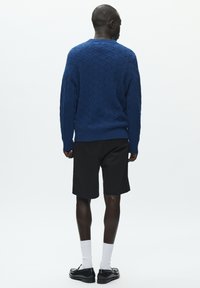 Pull tricoté bleu avec motif en losanges, poignets et ourlet côtelés, assorti à un short noir et des chaussures noires, vu de dos.