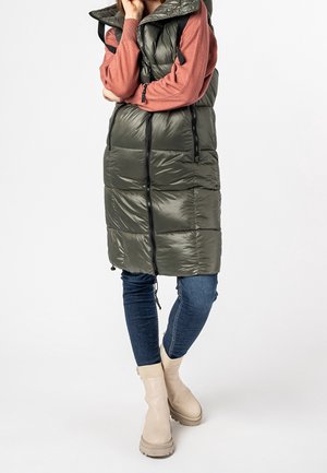 Personne portant un long gilet matelassé vert olive par-dessus un pull couleur rouille, un jean bleu et des bottines beige, debout les bras croisés.