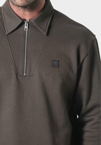 Pullover marrone con zip e colletto strutturato, caratterizzato da una discreta patch con logo e un tessuto liscio e testurizzato.