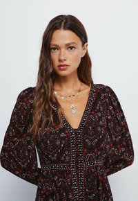 Černé a červené šaty se vzorem, nabíranými rukávy, hlubokým výstřihem do V a dekoračním prošíváním. Zlatý náhrdelník s kulatými přívěsky jako akcenty.