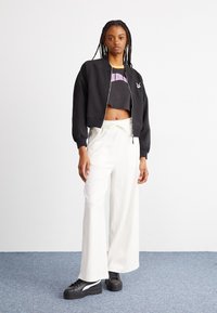 Veste bomber noire courte, pantalon large blanc avec des rayures verticales, et baskets noires avec des accents blancs. Le modèle pose devant un fond clair.
