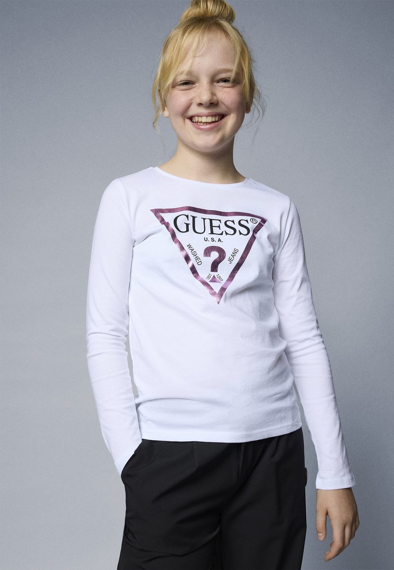 Smilende barn med blondt hår iført en hvit langermet skjorte med en stor fargerik Guess-logo, stående med hendene i lommene på svarte bukser.