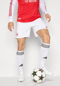 Rød trøje med hvide detaljer og teksten "FLY BETTER", hvide shorts og sort/hvide stribede sokker; står ved siden af en mønstret fodbold.