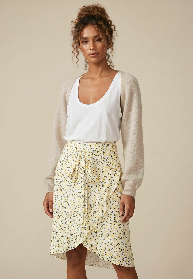 Femme aux cheveux bouclés porte un débardeur blanc, un cardigan beige et une jupe portefeuille jaune à fleurs sur fond beige uni.