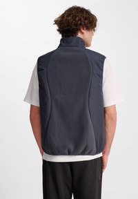 Mann in dunkelgrauem ärmellosem Fleece-Weste über einem weißen kurzärmligen Hemd, steht mit dem Rücken zur Kamera vor einem weißen Hintergrund.