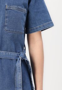Denim oblačilo z kratkimi rokavi, žepom na prsih in zavezanjem pasu. Barva je srednje modra s kontrastnim šivom.