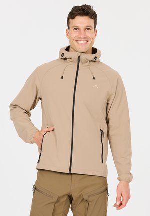 Lachende man die een beige capuchonjas en bruine cargobroek draagt, met één hand in de zak van de jas tegen een effen achtergrond.