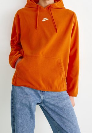 Personne portant un sweat à capuche orange Nike avec poche avant et un jean bleu clair, se tenant devant un fond blanc uni.