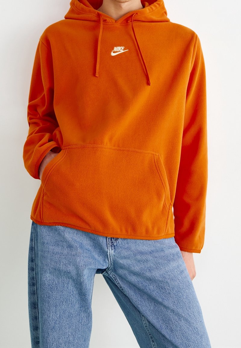 Person som bär en orange Nike-hoodie med framficka och ljusblå jeans, stående mot en slät vit bakgrund.