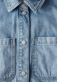 Camisa de mezclilla azul claro con cuello puntiagudo, botones de presión plateados, dos bolsillos en el pecho, costuras visibles y una tela suave y texturizada.