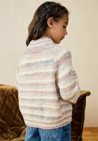 Neule, jossa on pehmeitä pastellisävyisiä raitoja, korkea kaulus, joustinneulereunus ja valuneet olkapäät, päällä siniset farkut.