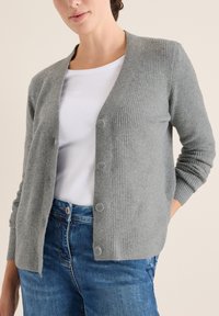 Grauer gerippter Cardigan mit langen Ärmeln und fünf Knöpfen vorne, getragen über einem weißen Hemd und blauen Jeans. Weiche Textur.