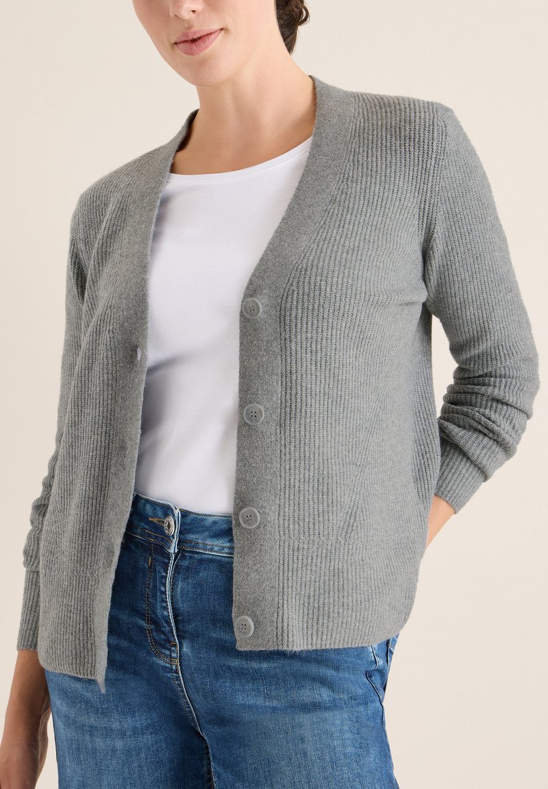 Grauer gerippter Cardigan mit langen Ärmeln und fünf Knöpfen vorne, getragen über einem weißen Hemd und blauen Jeans. Weiche Textur.